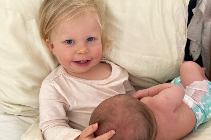 A little girl sitting down against a pillow, with her baby brother's head resting on her lap.