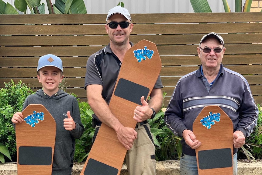 A boy on left, man in middle with sunglasses, an older man on right all holding large natural wood sandboards