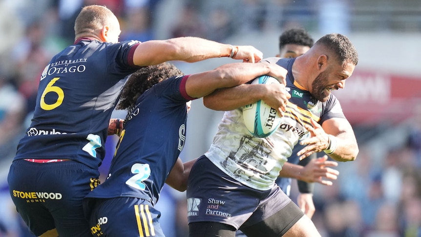 A Brumbies Super Rugby Pacific player holds the ball as he is tackled by Highlanders' players.