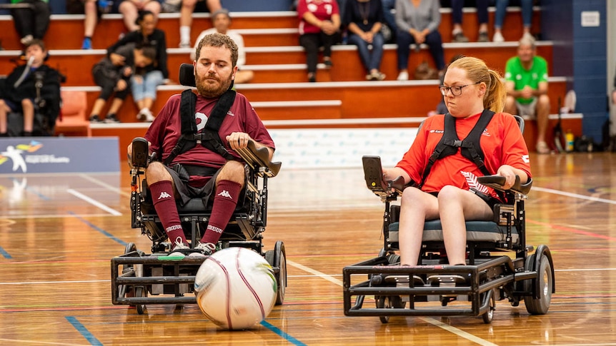 A man and a woman in electric wheelchairs battle for the ball with crowd in the background