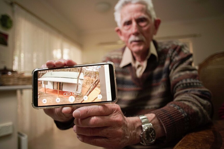 An elderly man holding a smartphone with a picture of a small house on it among trees