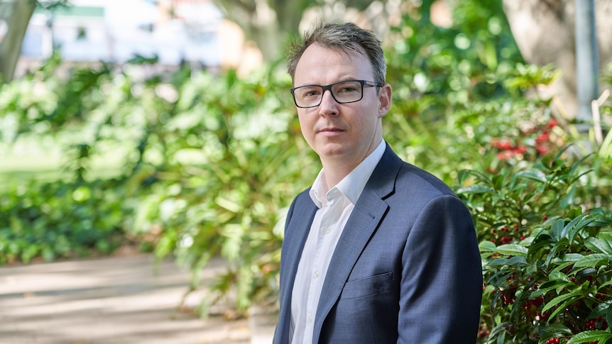 A man in a dark suit sits in front of some greenery.