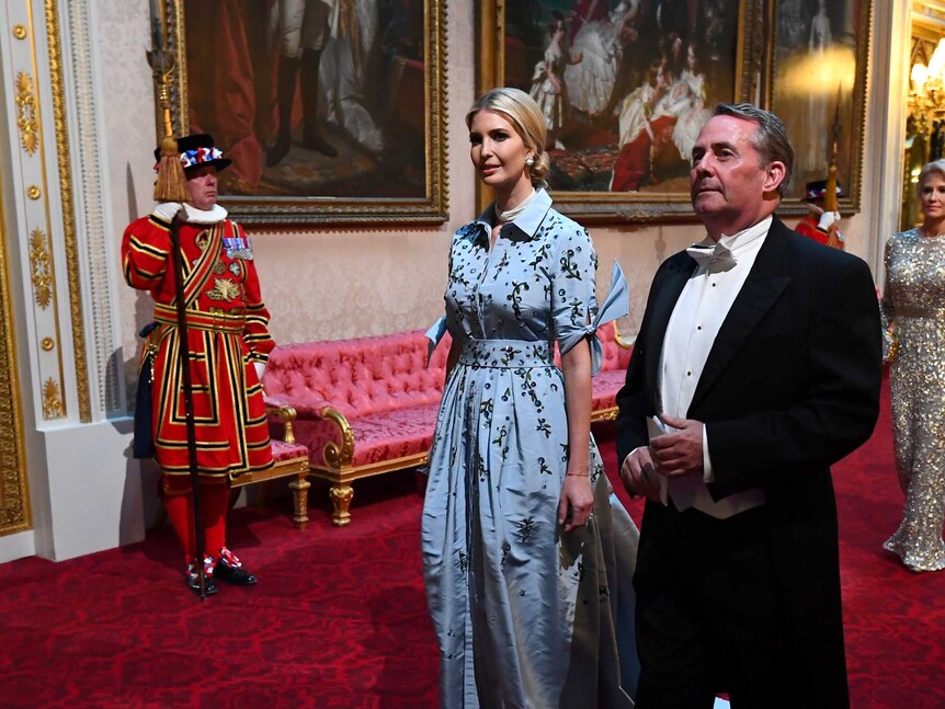 In an ornate corridor of Buckingham Palace, dignitaries in formalwear walk down bright red carpet in a rose pattern.