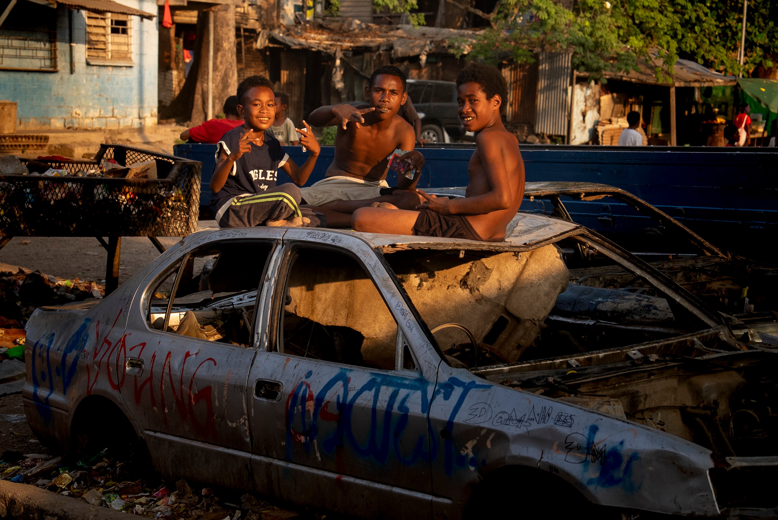 Three young boys sit on the sunken hood of a burnt-out car smiling at the camera