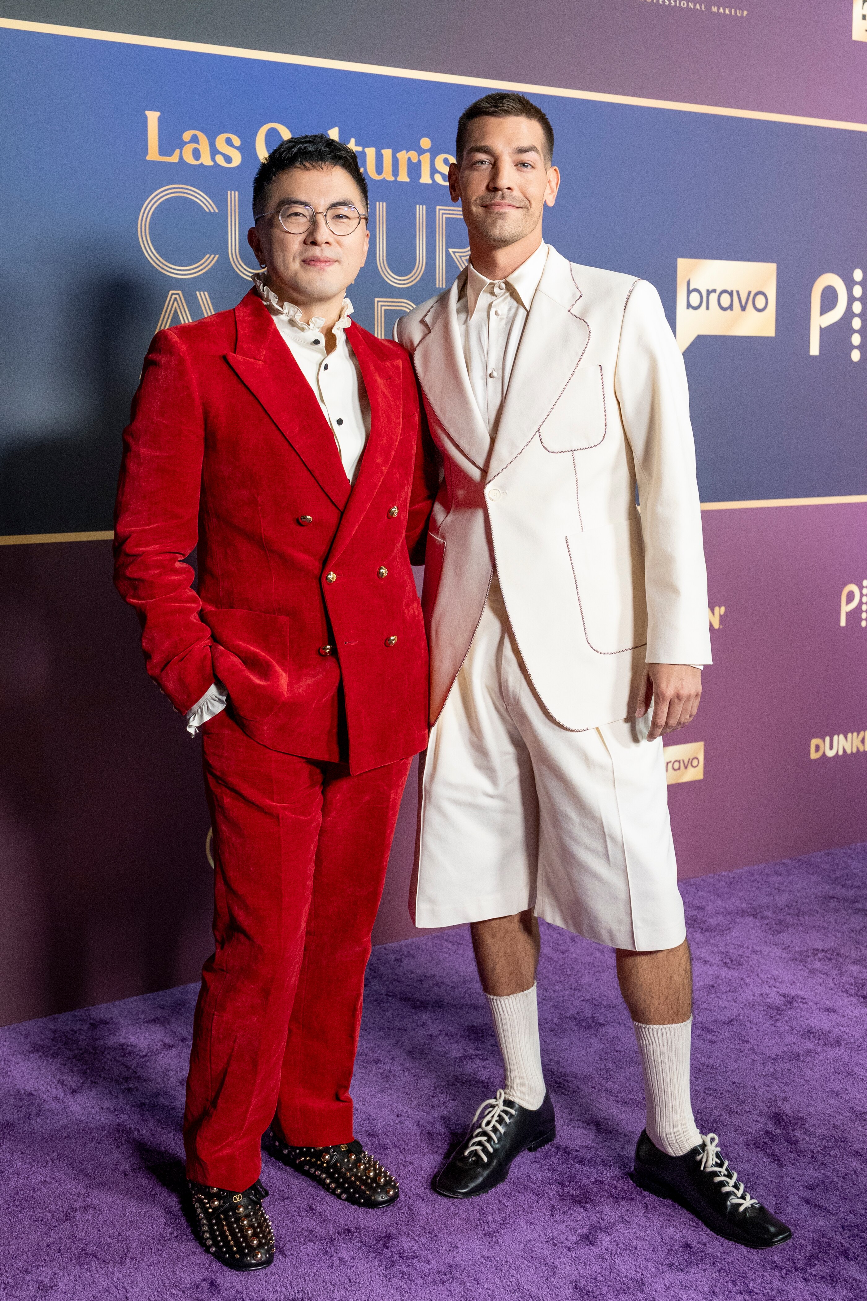 Two men pose together, one wears a red suit, the other in white, they're smiling at the camera