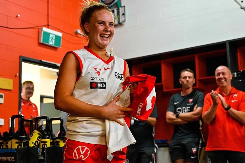 Ruby Sargent-Wilson smiles as she holds a swans guernsey in front of clapping club officials.