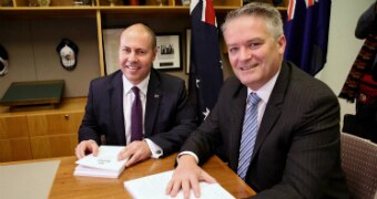 Treasurer Josh Frydenberg and Finance Minister Mathias Cormann smile at the camera while sitting at a desk with the Budget.