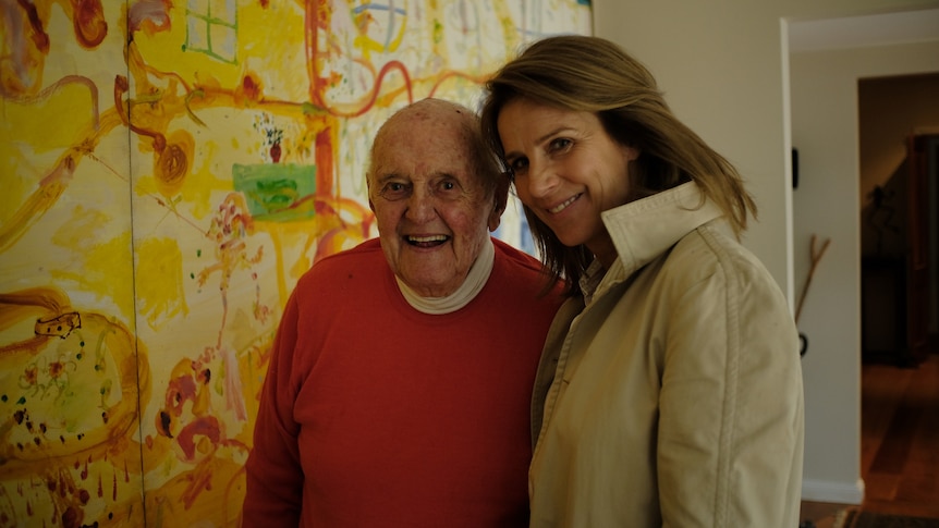 An elderly white man wearing a red jumper and a middle-aged white woman with brown hair stand beside a John Olsen painting