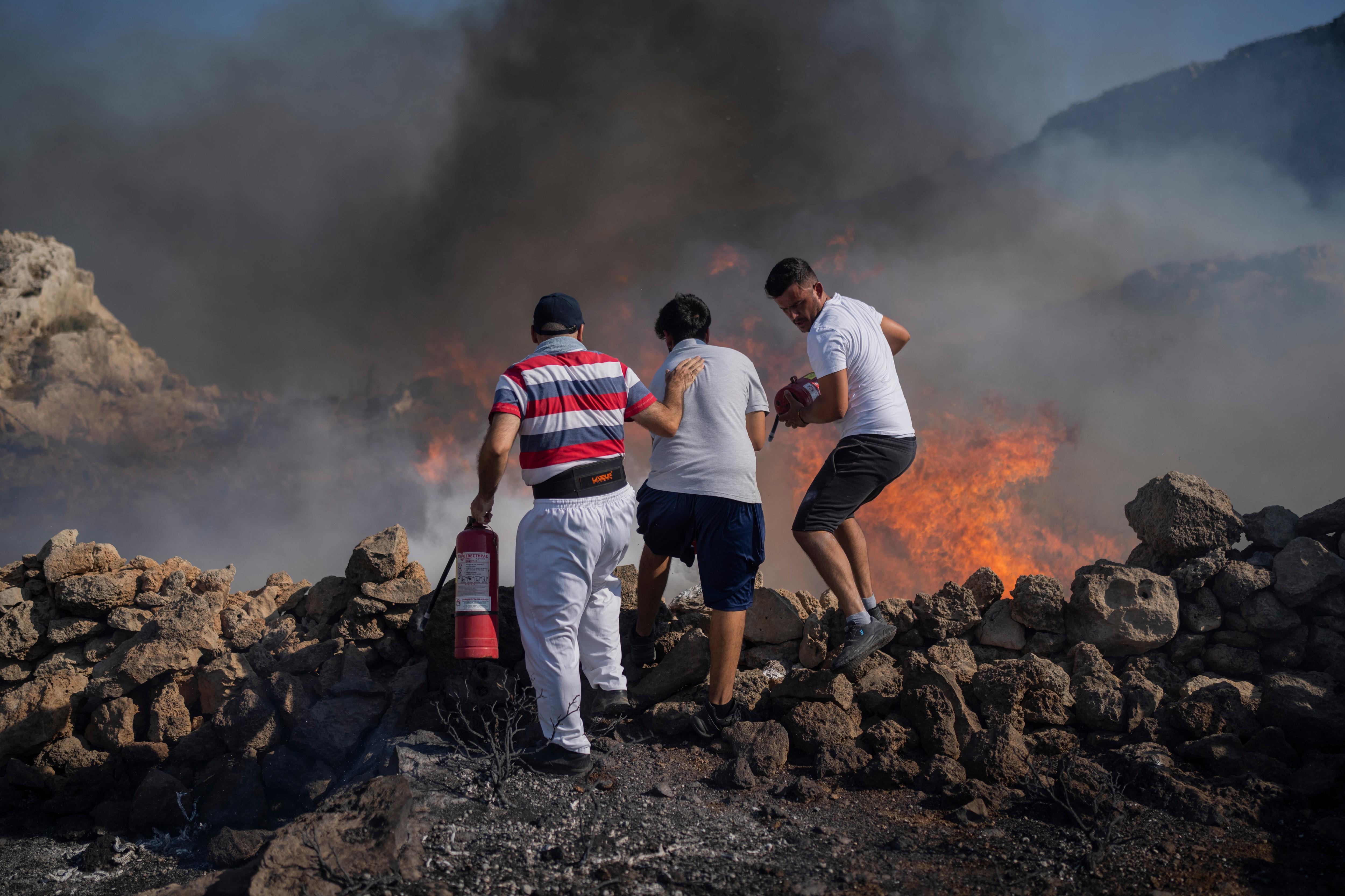 Three men stand at the edge of a rock face using extinguishers to try and extinguish a large fire with angry flames