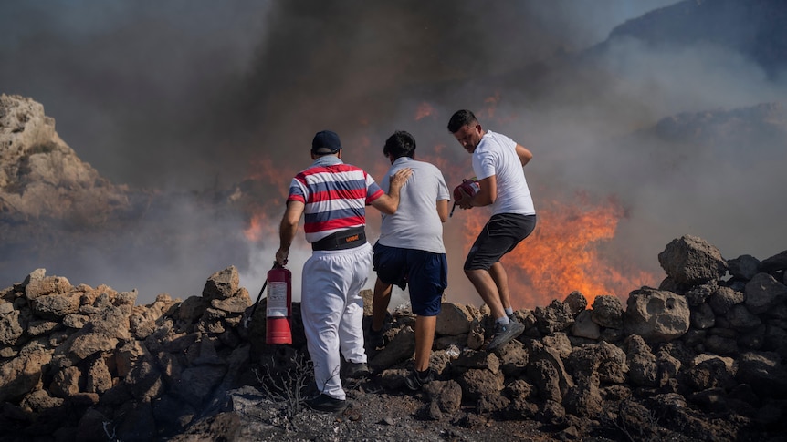 Three men stand at the edge of a rock face using extinguishers to try and extinguish a large fire with angry flames