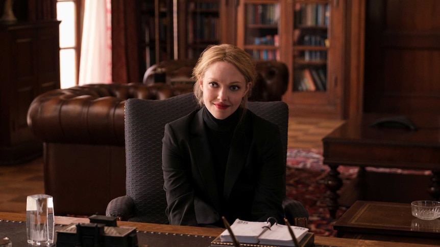A blonde woman sits at a large desk in an opulent office.