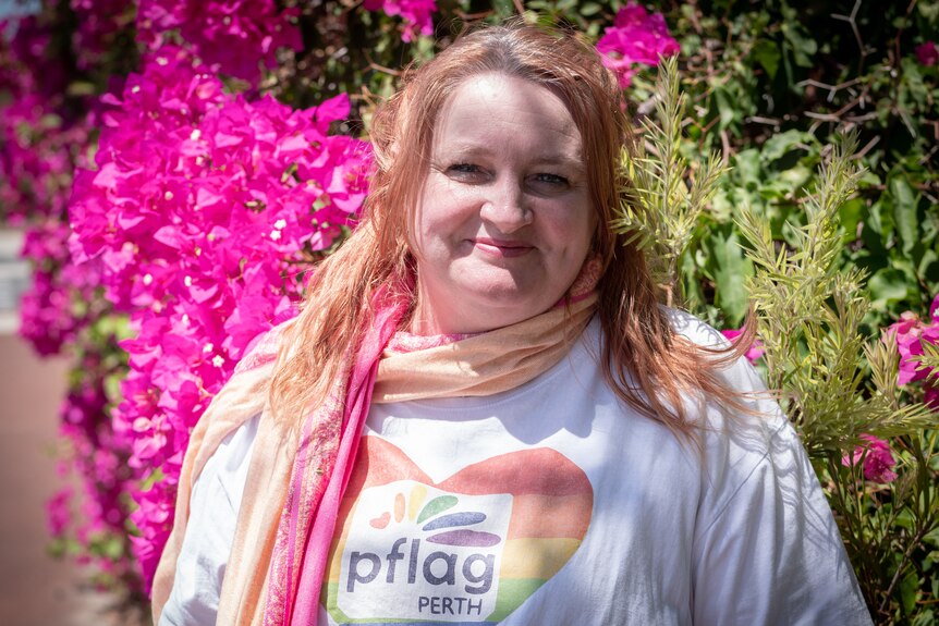 Woman with red hair and pflag t-shirt stands in front of pink plant, smiling