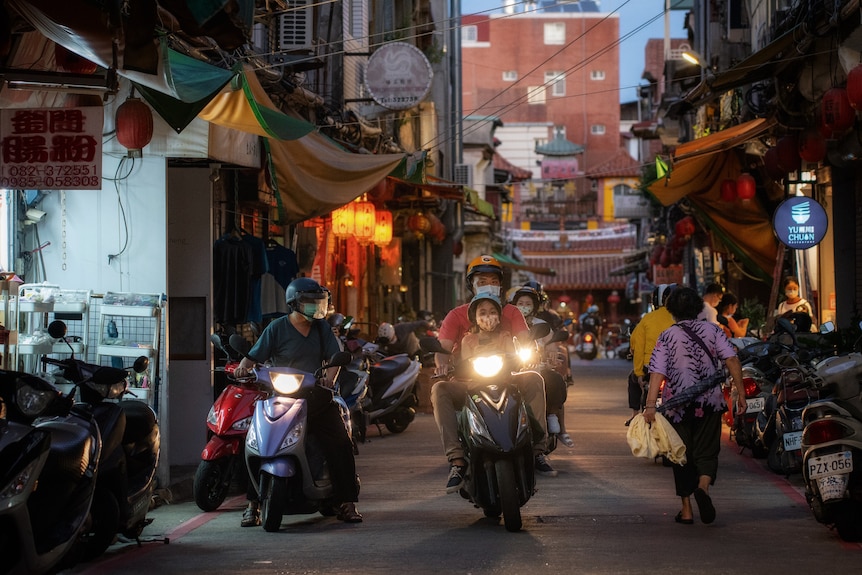 Two people with passengers at the back drive down a narrow, busy alleyway on a mortorbike