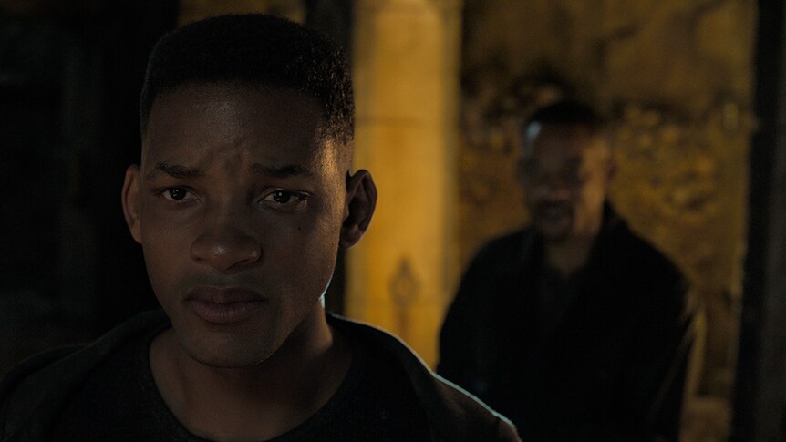 An unknown man stands behind Will Smith (left) with a sad expression, in a shadowy and dimly lit space.