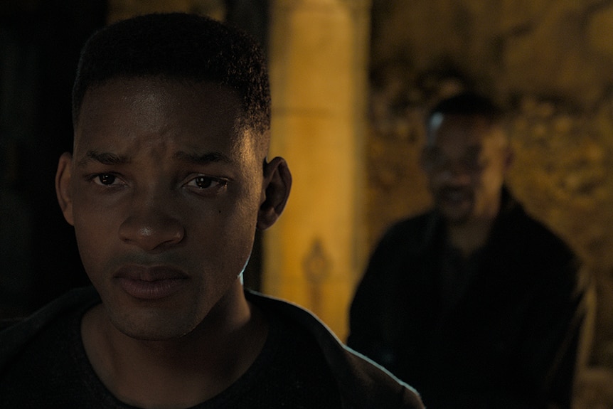 An unknown man stands behind Will Smith (left) with a sad expression, in a shadowy and dimly lit space.