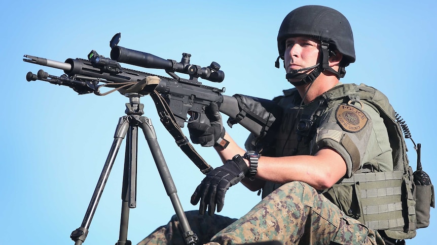 Policeman dress in army fatigues sits with a large black gun on a tripod.