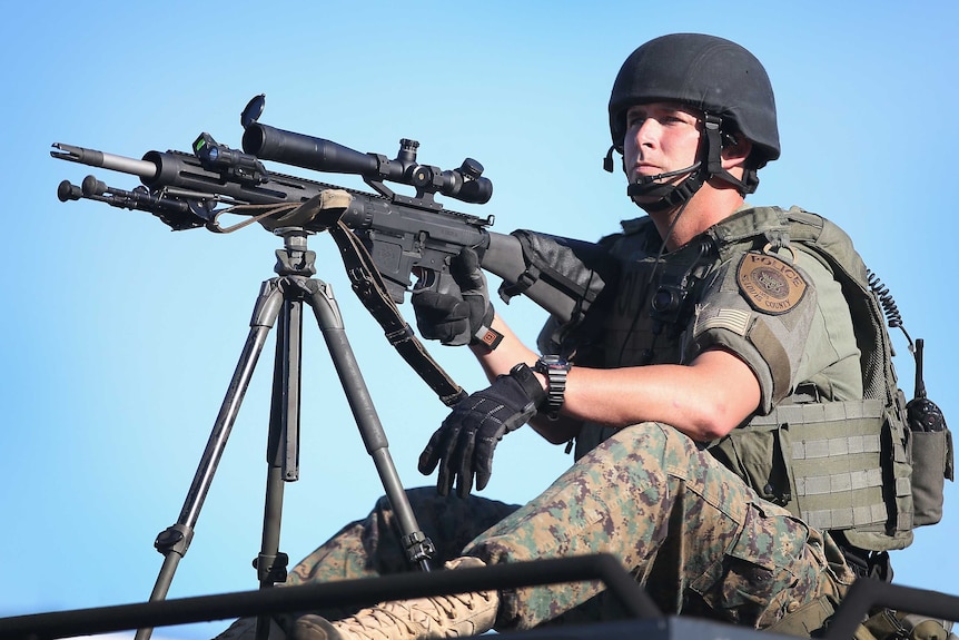 Policeman dress in army fatigues sits with a large black gun on a tripod.