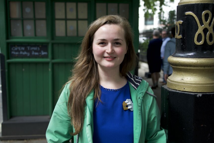 A young woman stands in front of a small, green, heritage-listed cab shed in London.