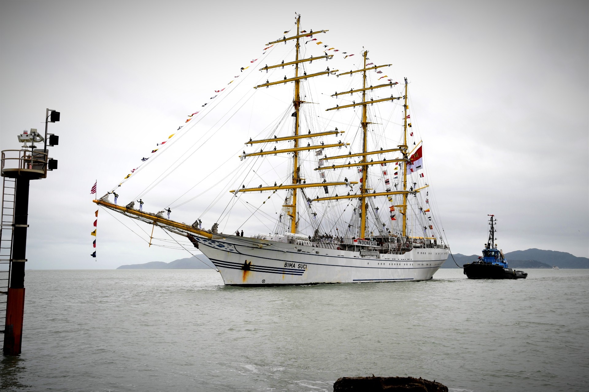 A large sailing boat with people sitting on the mast sails with grey skies behind