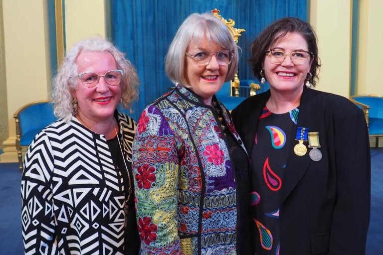 Three woman smile at the camera in front of a stage with a blue curtain.