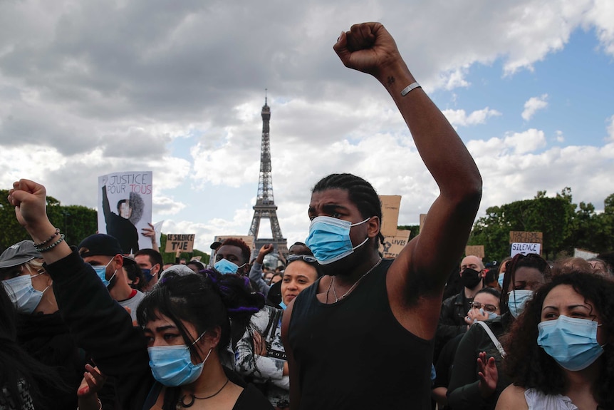 A man wearing a mask raises his fist in protest. Behind him is a crowd and the Eiffel Tower.