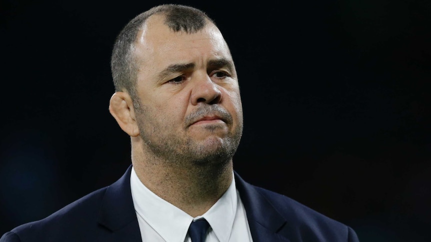 Michael Cheika, wearing a dark suit, frowns as he stands in front of a dark background.