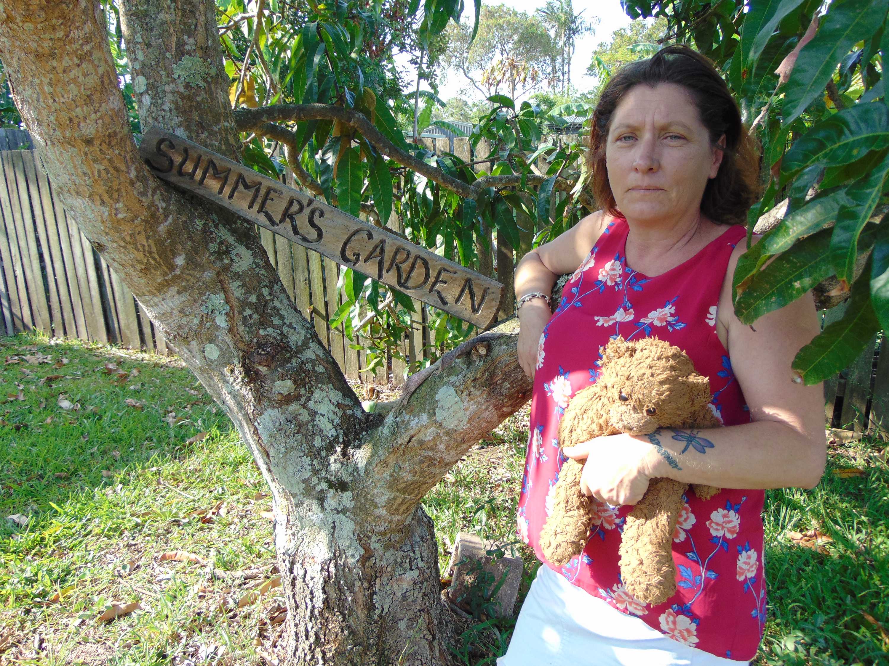 Middle-aged woman holding teddy bear and leaning against a mango tree with the sign "Summer's Garden".
