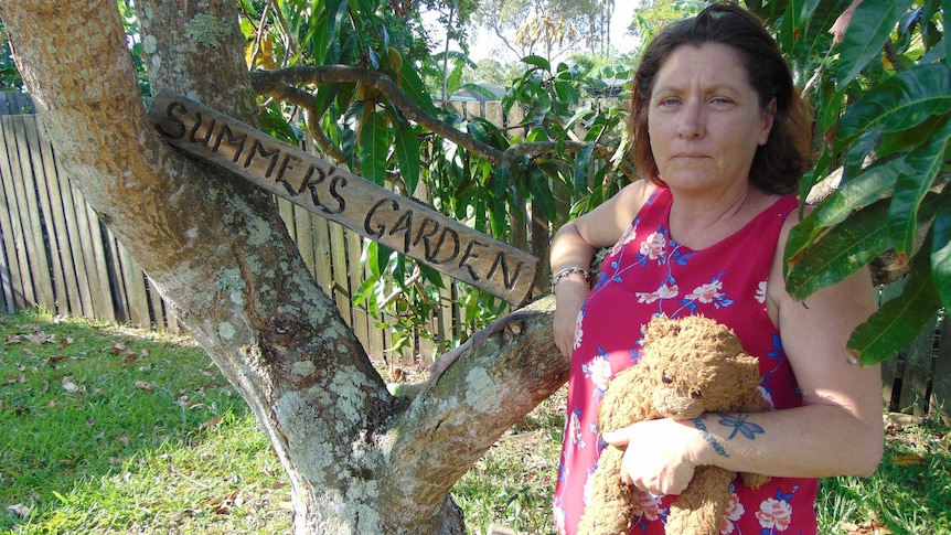 Middle-aged woman holding teddybear standing next to mango tree with sign, Summer's Garden.