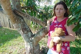 Middle-aged woman holding teddy bear and leaning against a mango tree with the sign "Summer's Garden".