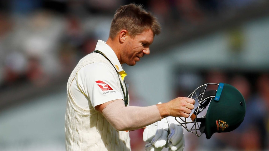 A male cricketer walking while holding his helmet in his left hand and gloves in his right, as he looks towards the ground.