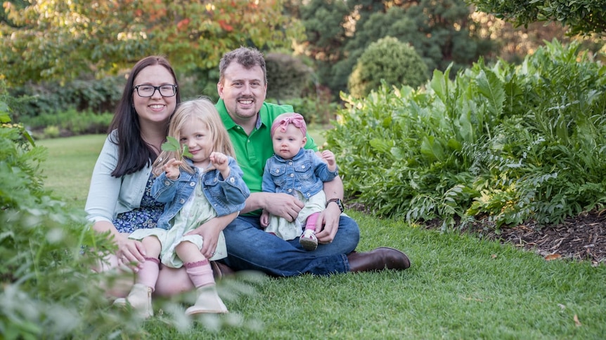 A woman with a toddler on her lap and a man with a baby on his lap sit side by side on the grass smiling.