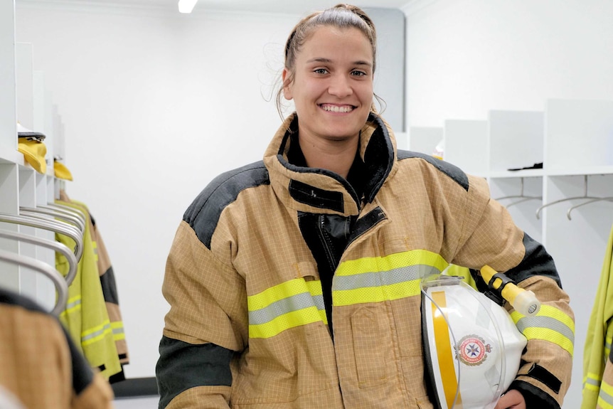 A woman in a firefighting uniform holds a helmet under her arm, smiling in the locker room.