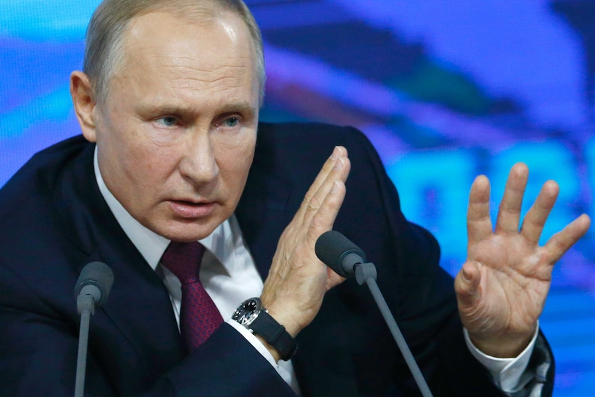 Vladimir Putin holds up his hands while speaking at a lectern