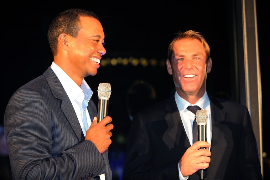Tiger Woods and Shane Warne smile holding microphones at an event.