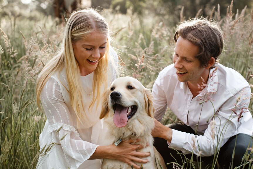 Brooke Stratton and her partner Nathan pat their golden retriever in long grass.