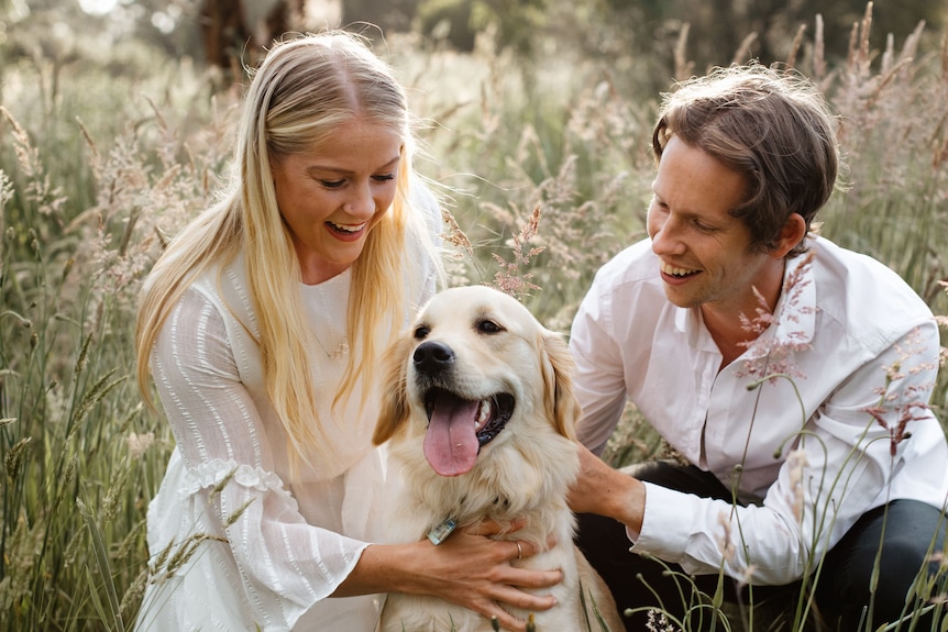 Brooke Stratton and her partner Nathan pat their golden retriever in long grass.