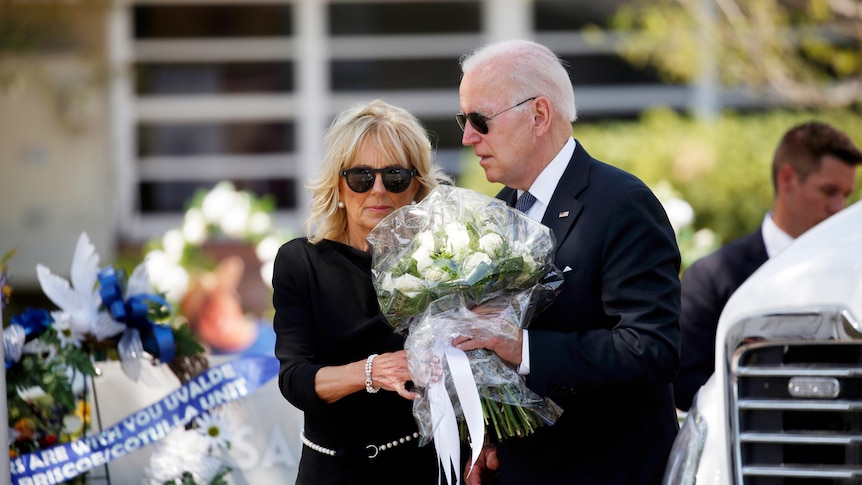An elderly white couple wearing dark formal clothes and sunglasses holds a large bouquet of flowers