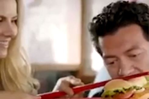 A blond-haired woman holding a burger up to a man's face with giant red chopsticks as he tries to eat it