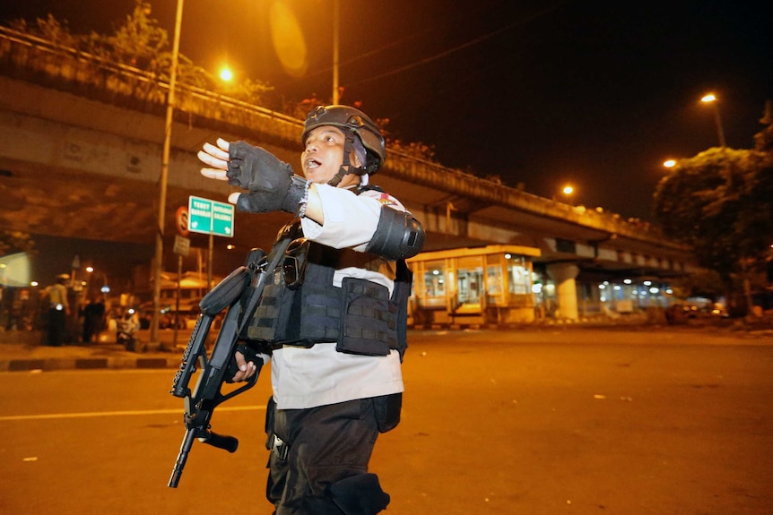 A police officer with a high-powered gun gestures at the scene of an explosion.