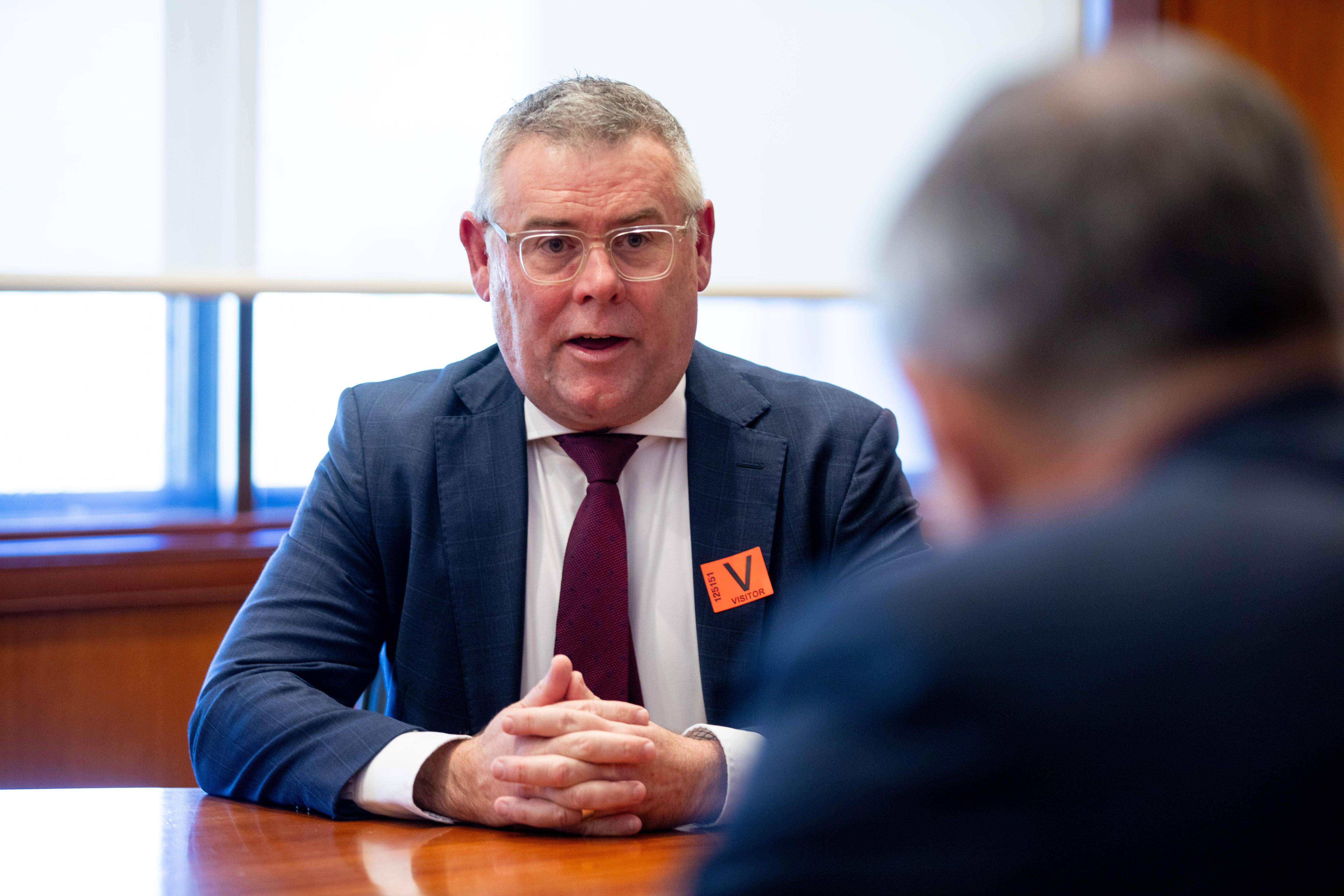 Murray Watt seated in front of a window, hands clasped, with the back of a head in the foreground.
