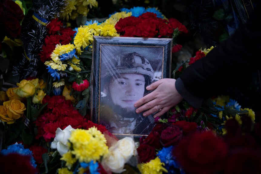 a photo of an Ukrainian soldier sits in a picture frame surrounded by flowers as a woman touches the photo