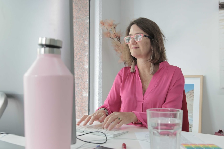 A woman wearing a pink shirt and glasses sits at her computer.