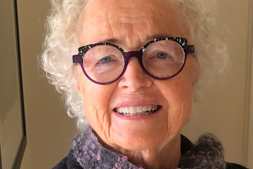 A 75-year-old woman with white hair and glasses, smiling while posing for a photograph.