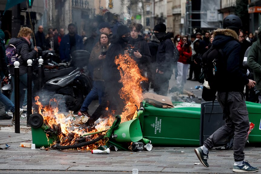 A rubbish bin on fire in the middle of a street surrounded by people.