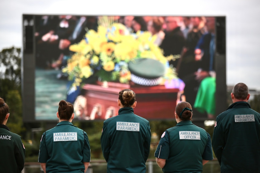 A row of ambulance officers in uniform watching a funeral on a large outdoor screen.