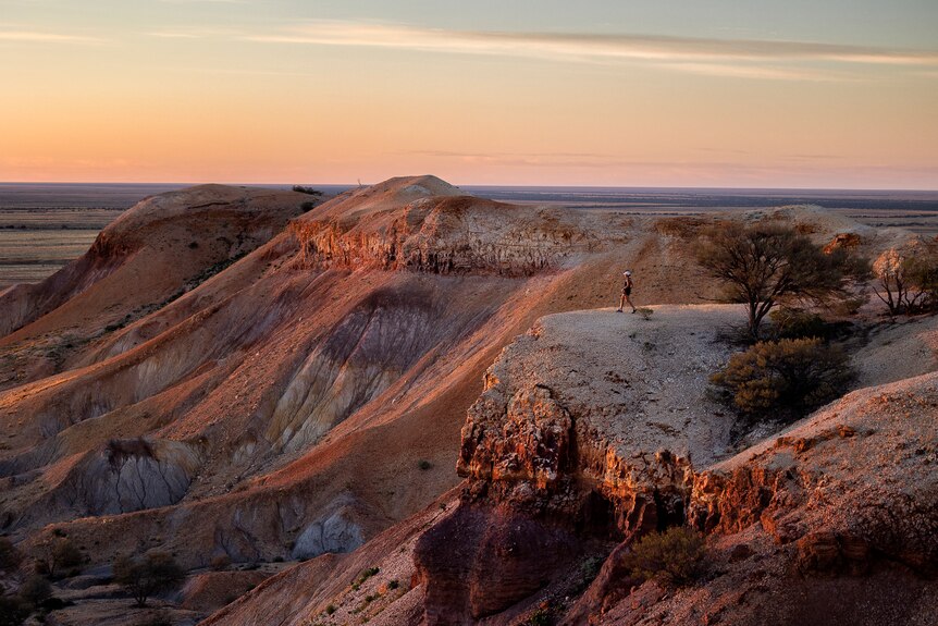 A woman stands on the edge of red cliffs with the sun rising over them, a desert extends beyond. 