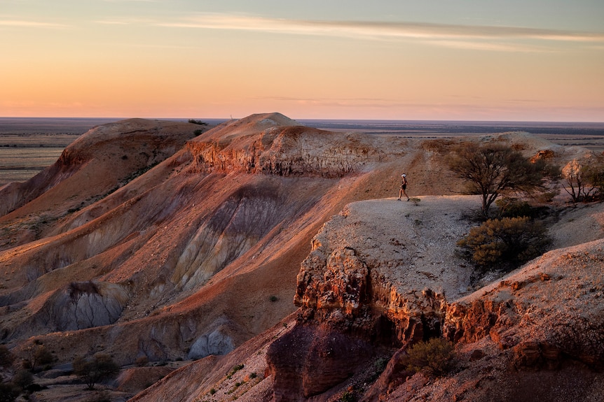 A woman stands on the edge of red cliffs with the sun rising over them, a desert extends beyond. 