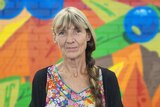 A woman with a bright floral dress and black cardigan stands in front of a brightly coloured mural.