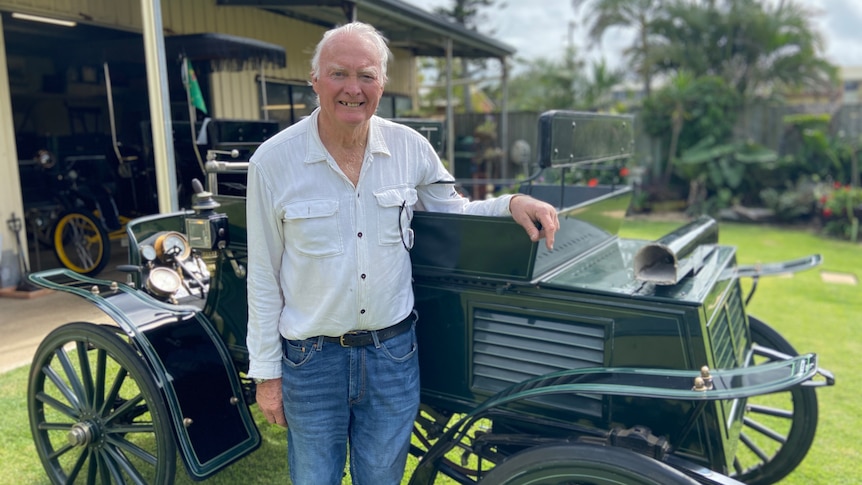 An elderly man standing beside a forest green vintage car in his backyard