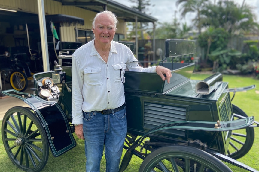 An elderly man standing beside a forest green vintage car in his backyard
